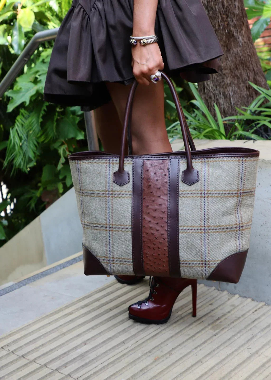 Person holding a plaid tote bag with brown leather straps outdoors.
