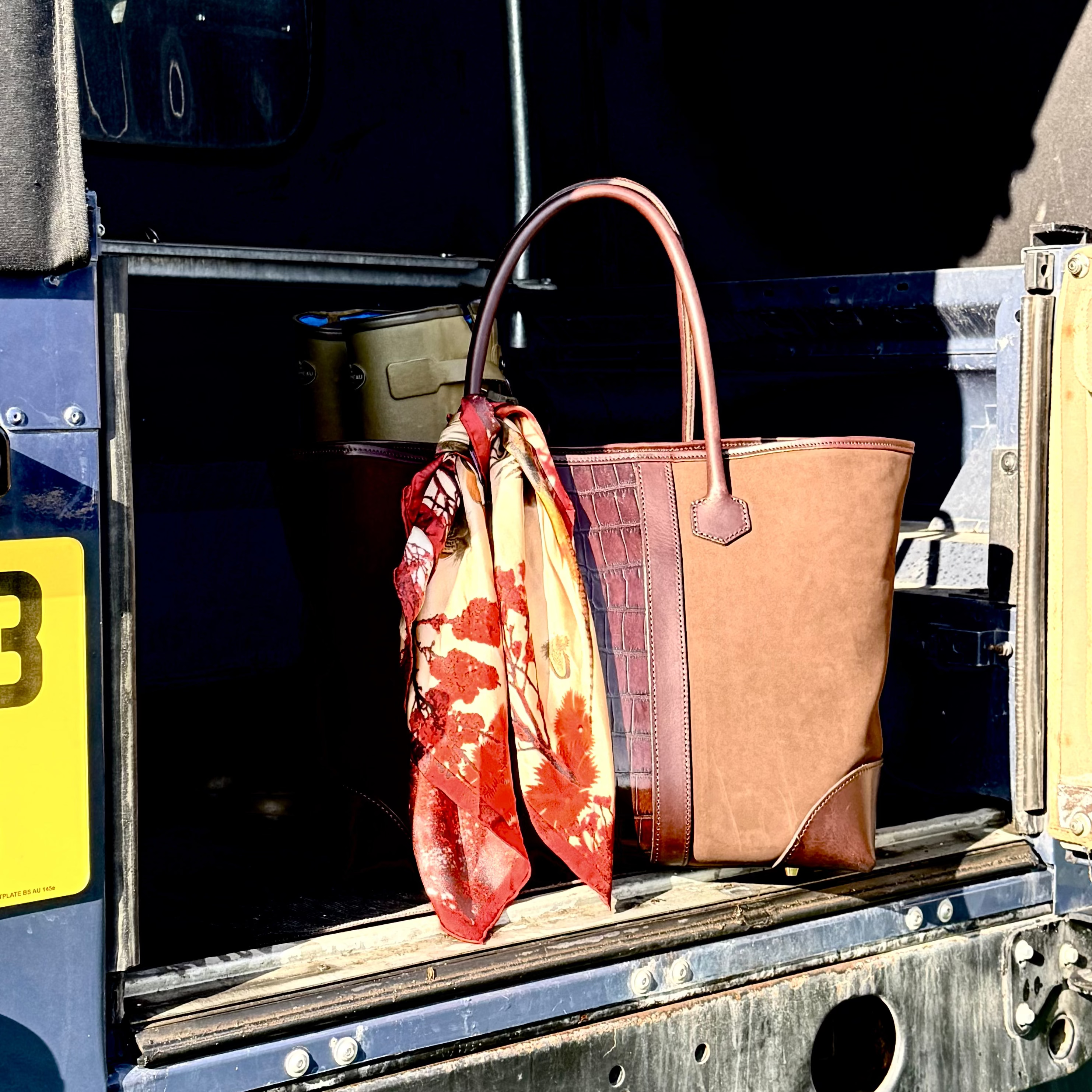 Brown handbag with a floral scarf draped over it, placed in the open back of a Range Rover