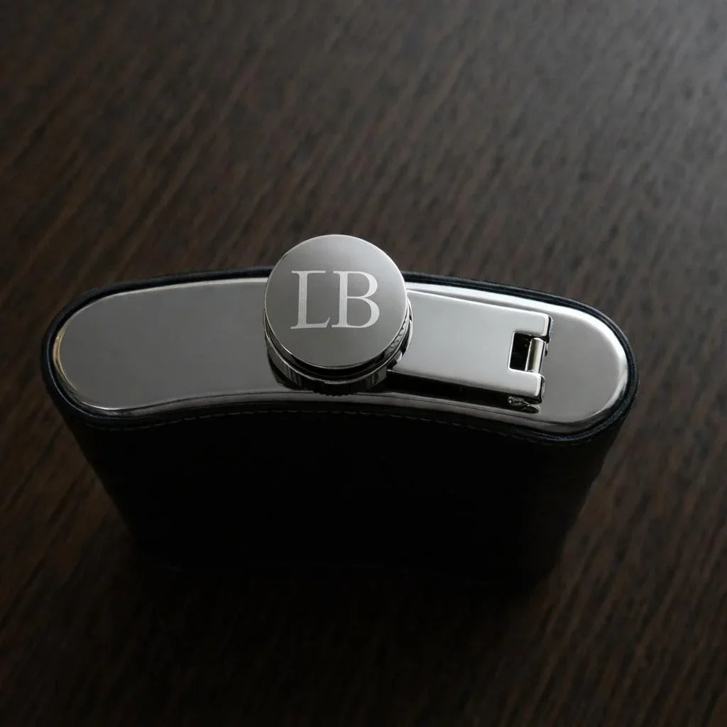 Black hip flask with a metallic cap featuring initials on a dark wooden surface
