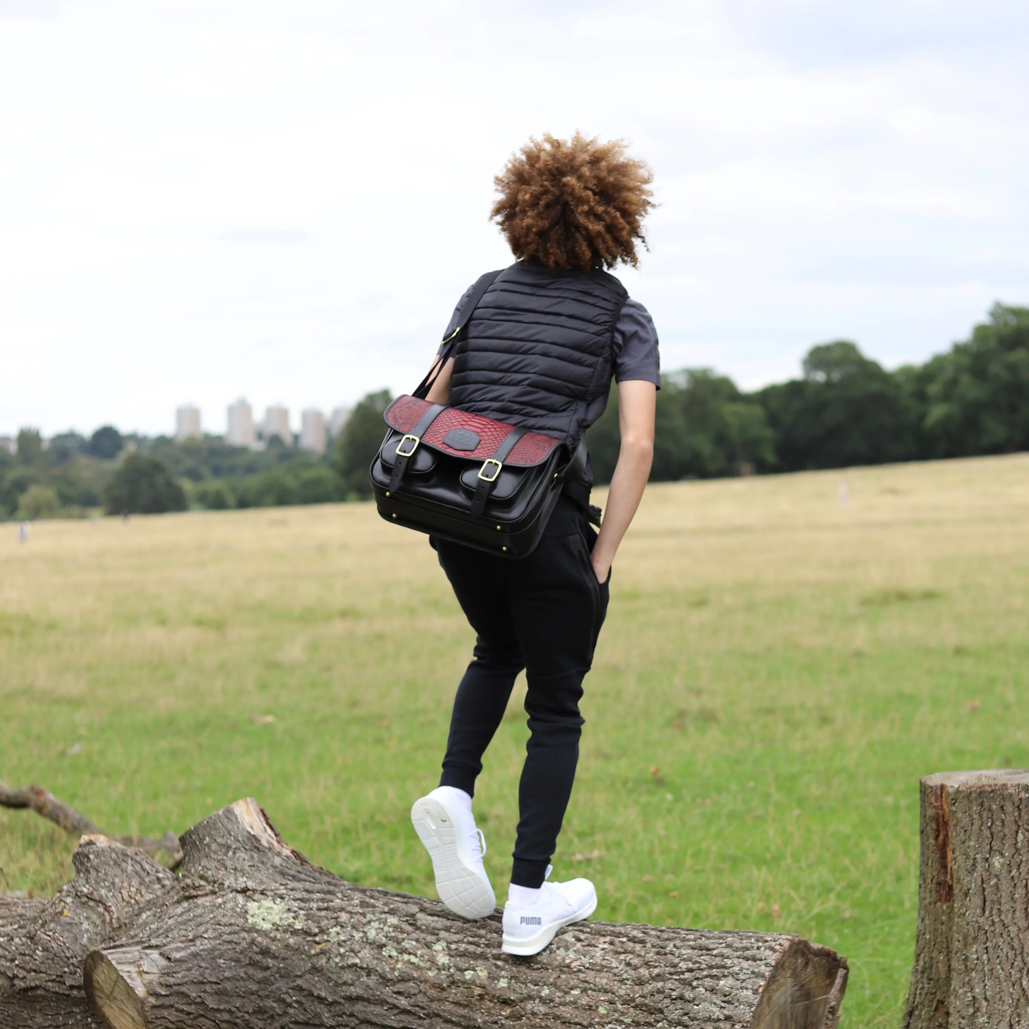 HERA-CELENE BLACK WITH BURGUNDY SNAKE CARTRIDGE BAG BEING WORN BY A PERSON STANDING ON A LOG IN THE MIDDLE OF A FIELD