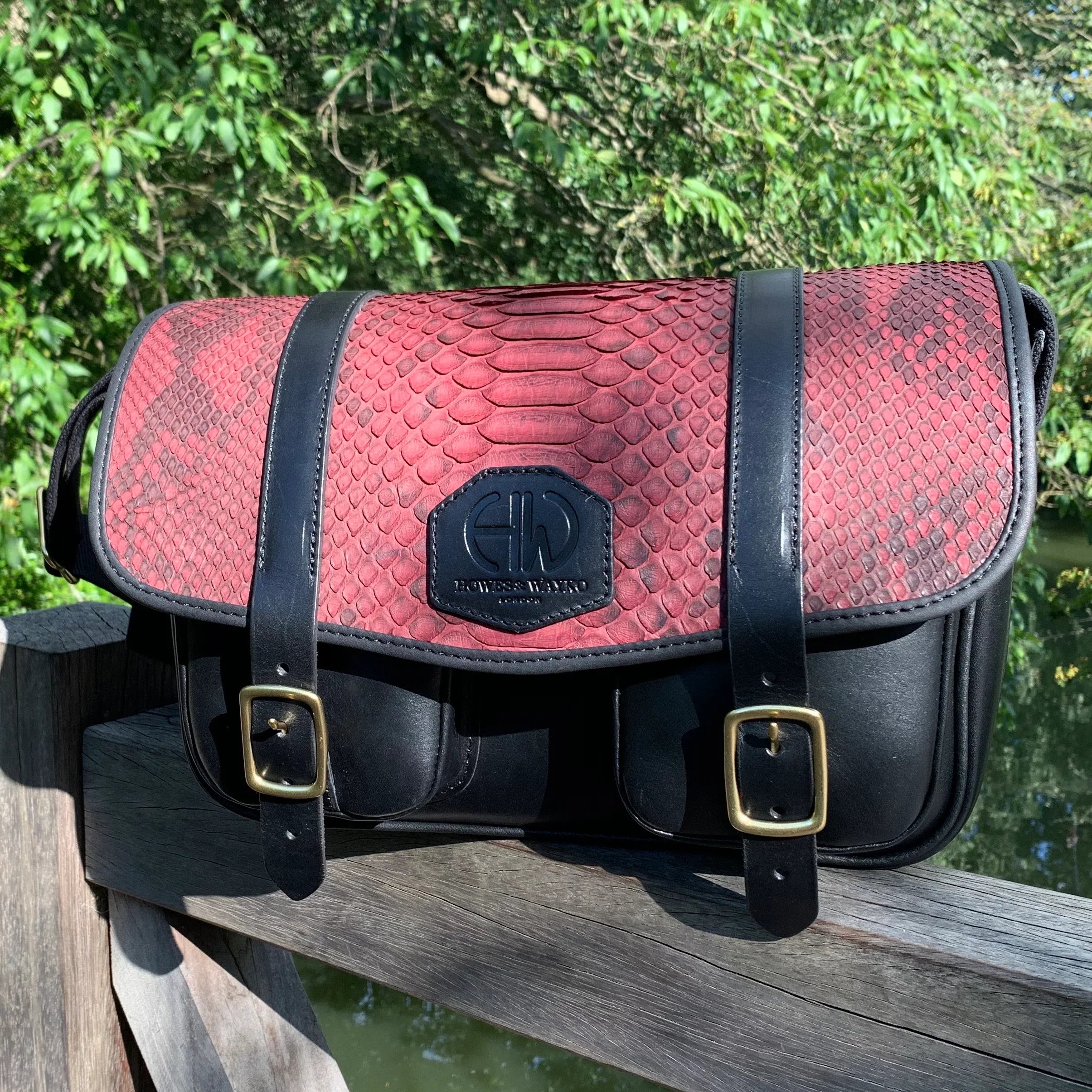 HERA-CELENE BLACK WITH BURGUNDY SNAKE CARTRIDGE BAG front view of bag perched on a wooden fence with trees in the background