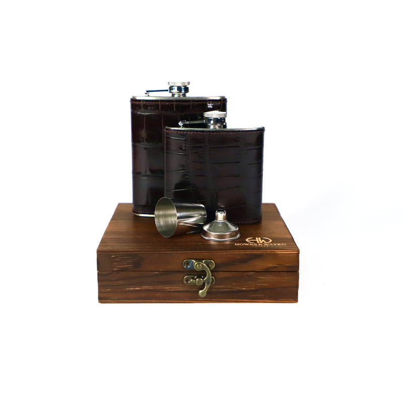 Two dark moc-croc leather flasks with silver caps on a wooden stand with a white background