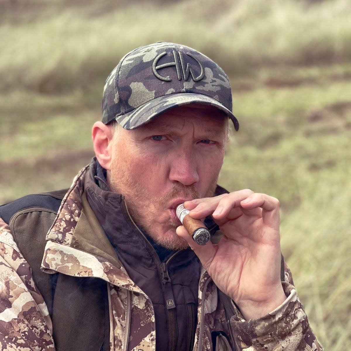 A man in camouflage clothing and a Howes and Wayko camouflage cap smoking a cigar outdoors of Africa
