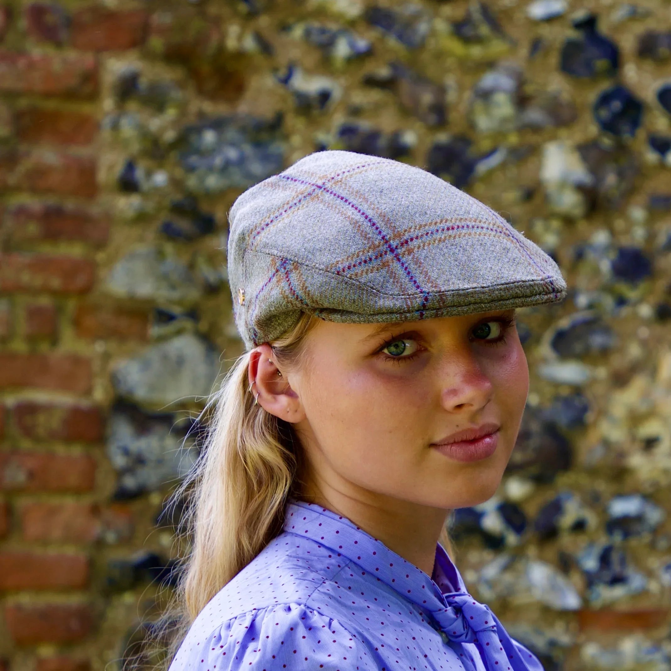 Woman wearing a checkered tweed style flat cap against a textured wall.