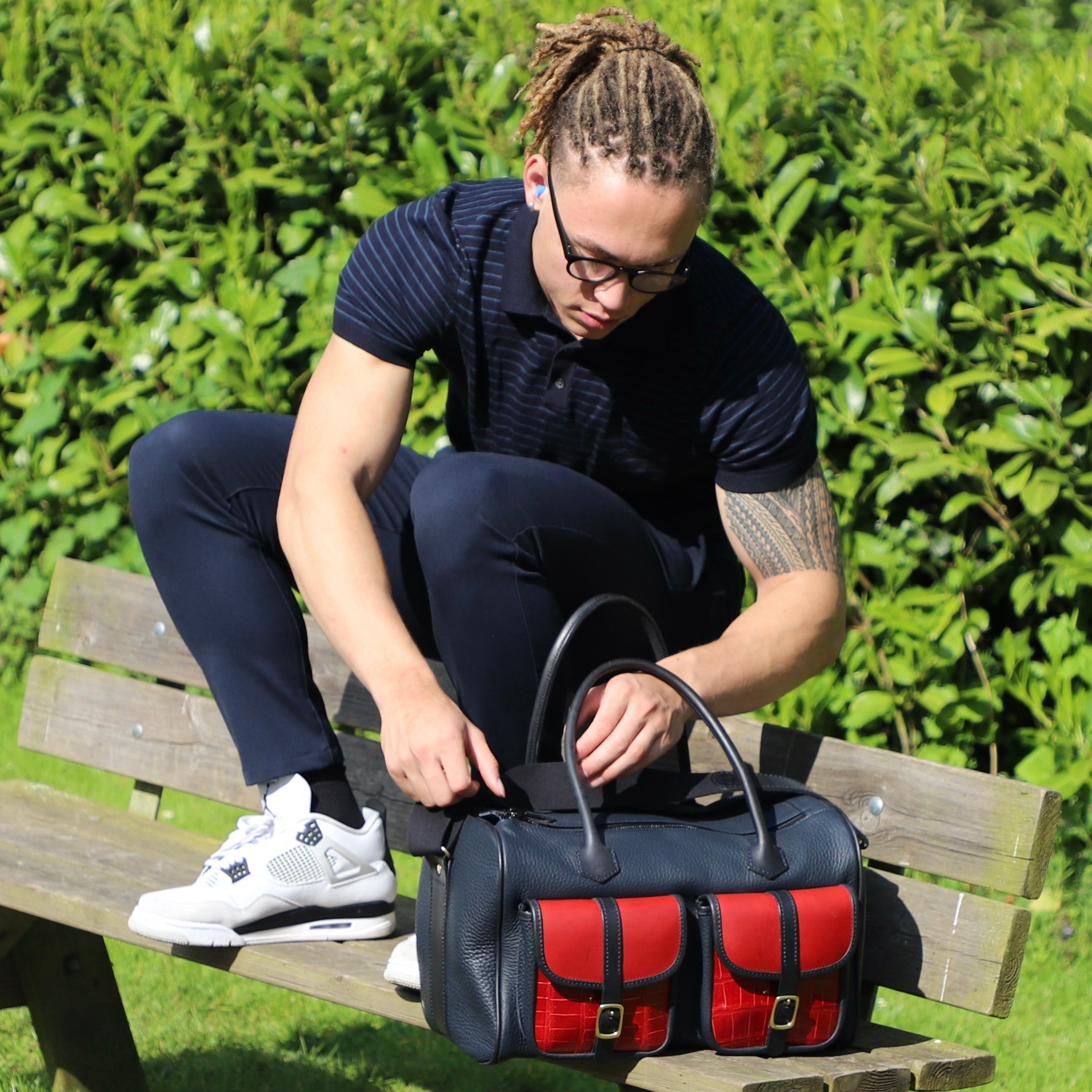 a man with a leather range bag in the erin collection sitting on a bench with the range bag about to unzip the bag, bushes and greenery in the outdoor background