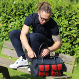 a man with a leather range bag in the erin collection sitting on a bench with the range bag about to unzip the bag, bushes and greenery in the outdoor background